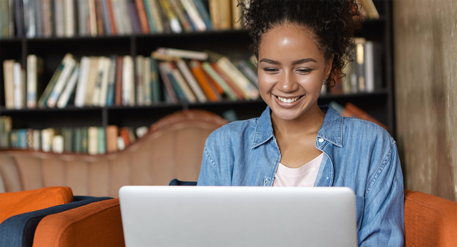 Happy Black young woman in jean shirt sitting at sofa using laptop.