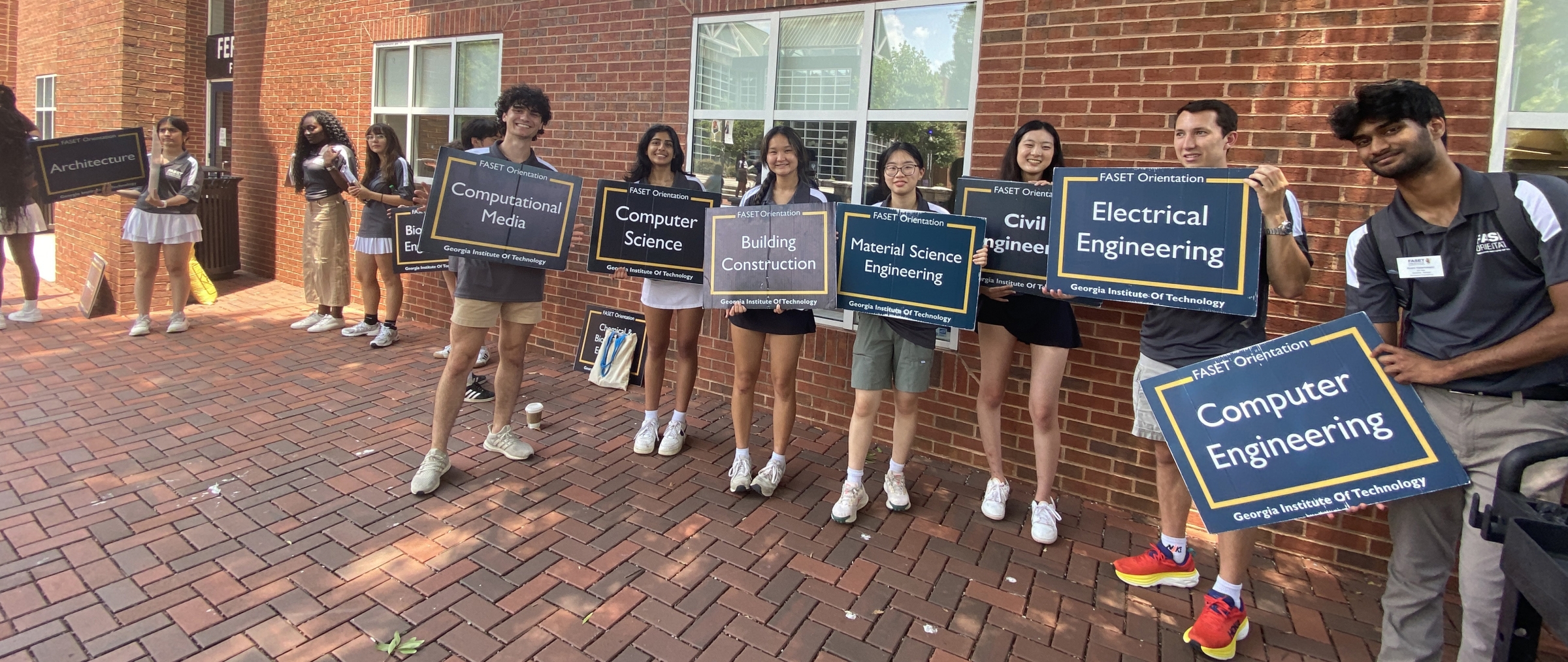A group of seven college students hold signs displaying college names, smiling and leaning in